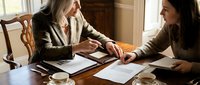 Two professional businesswomen collaborating on documents and contracts in elegant office setting with warm natural lighting and coffee cups.