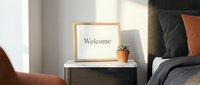 Cozy modern bedroom with a wooden framed welcome sign and small succulent on a marble nightstand beside a bed with gray and orange pillows.