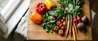Fresh organic vegetables and herbs arranged on rustic wooden cutting board in bright kitchen. Colorful produce including tomatoes, peppers, leafy greens for healthy cooking.