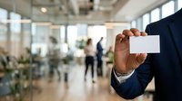 Professional man in dark suit holding blank white business card in modern office lobby with colleagues in background.