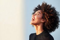 Young African American woman with natural curly hair laughing joyfully against soft background, wearing black top, side profile showing genuine happiness and freedom.