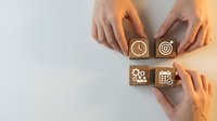 Hands arranging wooden cubes with business icons including clock, target, gear, and calendar on light background for planning concept.