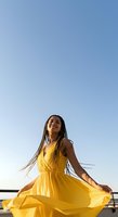 Young woman with long braided hair and medium brown skin tone wearing a flowing yellow halter dress, smiling with eyes closed against clear blue sky, outdoor lifestyle and fashion portrait.