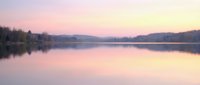Serene lake at dawn with pink and golden sky reflecting on calm water, surrounded by bare trees and distant mountains, peaceful nature landscape.