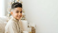 Young boy with dark hair and light skin tone, wearing a beige button-up shirt, smiling at camera with blurred holiday decorations in background.