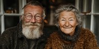Happy senior Caucasian couple smiling warmly at camera in cozy home setting with soft lighting and comfortable sweaters for lifestyle content.
