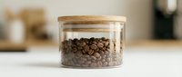 Roasted coffee beans stored in a round glass jar with a wooden lid on a white kitchen counter, soft blurred background.