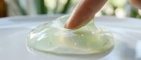 Finger touching translucent gel substance with glossy texture on white surface, demonstrating skincare product consistency for cosmetic and beauty applications.