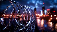 Razor wire fence at night with blurred city lights creating atmospheric urban security scene. Military or prison perimeter with bokeh effect in background.