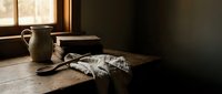 Vintage rustic kitchen scene with ceramic pitcher, wooden cutting board and linen cloth on weathered table near window with warm natural lighting.