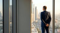 Confident businessman in a navy suit standing at a floor-to-ceiling office window overlooking a modern city skyline at sunset.