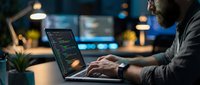 Male programmer coding on laptop in modern office environment with bokeh lights and multiple monitors creating atmospheric workspace for software development.