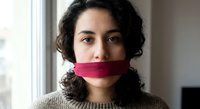 Young Middle Eastern woman with curly hair and pink tape over mouth in domestic interior representing censorship and silence concepts.