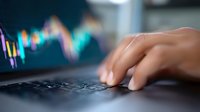 Close-up of hands typing on laptop keyboard with blurred financial trading chart in background, showing stock market data analysis and investment monitoring.