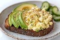 Healthy avocado toast with scrambled eggs on dark bread served with cucumber slices on ceramic plate for nutritious breakfast or brunch meal.
