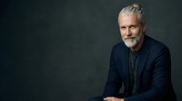 Mature man with gray hair and beard wearing dark blazer and shirt against neutral background, professional portrait for business and corporate use.