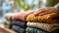 Stack of folded colorful knitted sweaters with woman's hand touching soft mustard yellow garment. Cozy autumn winter clothing collection.