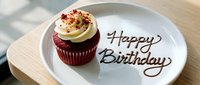 Red velvet cupcake with cream frosting on a white plate with Happy Birthday written in chocolate sauce, served on a wooden tray.