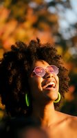 Joyful African American woman with curly hair wearing pink sunglasses laughing outdoors in warm autumn sunlight with colorful fall foliage background.