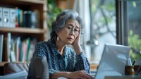 Tired senior Asian woman looking stressed while working from home on digital tablet, expressing fatigue and exhaustion during remote work in cozy home office setting.