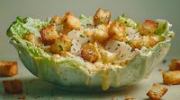 Fresh caesar salad in iceberg lettuce bowl topped with golden croutons and parmesan cheese, creamy dressing drizzled over crisp greens on green background.