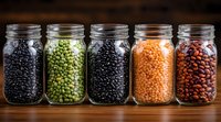 Various dried legumes and pulses in glass mason jars on wooden surface including black beans, green peas, lentils and kidney beans. Food storage concept.