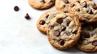 Fresh baked chocolate chip cookies with crispy edges and soft centers scattered on white surface, close up view showing gooey texture and melted chips.