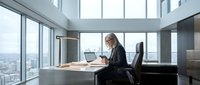 Young woman with long dark hair, South Asian ethnicity, wearing professional black blazer, working at wooden desk with laptop in modern high-rise office with floor-to-ceiling windows overlooking cityscape. Corporate workspace concept.