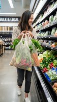 Young woman of East Asian descent holding reusable mesh shopping bags filled with fresh vegetables in grocery store produce section, sustainable shopping and eco-friendly lifestyle.