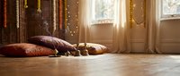 Cozy bohemian bedroom interior with decorative pillows on wooden floor, warm sunlight streaming through window with fairy lights creating ambient atmosphere.