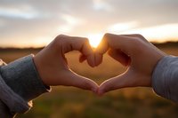 Hands forming heart shape against golden sunset sky creating romantic silhouette with warm sunlight streaming through fingers in peaceful outdoor setting.