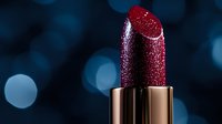 Glittering dark red lipstick with metallic gold case against blurred blue bokeh background. Luxury cosmetic product photography for beauty and glamour advertising.