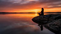 Woman in silhouette sitting on rocky outcrop meditating at golden sunset over calm lake water, peaceful nature scene for wellness and mindfulness content.