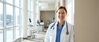 Confident female doctor in white coat with stethoscope smiling in modern hospital corridor. Professional healthcare worker portrait for medical marketing.