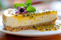 Slice of passion fruit cheesecake with graham cracker crust, topped with fresh passion fruit pulp, seeds, purple edible flower and mint leaf garnish on white plate.