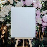 Blank white canvas on wooden easel with pink and white floral backdrop for wedding signage, event displays, and romantic celebration designs.