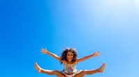 Young girl with curly brown hair and medium skin tone jumping joyfully against clear blue sky, arms spread wide, wearing colorful casual clothing, expressing happiness and freedom outdoors.