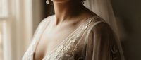 Young South Asian woman in elegant beige embroidered dress standing by window with soft natural lighting creating warm atmospheric portrait for fashion.