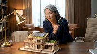 Mature woman with gray hair and light skin tone, wearing dark blazer, examining architectural model of residential house on wooden desk in modern office with brass lamp and city view for real estate, architecture, and business concepts.