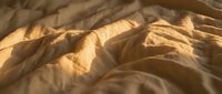 Unmade bed with golden sunlight casting shadows across crumpled beige linen sheets, creating texture and depth for bedroom, interior design, and lifestyle content.