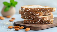 Fresh sliced whole grain bread with sesame seeds stacked on wooden cutting board, scattered almonds and green plant decoration in background.
