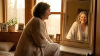 Middle-aged woman with curly hair in cozy sweater looking at herself in bedroom mirror with warm natural lighting creating peaceful morning self-care moment.