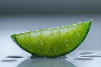 Fresh juicy lime wedge with water droplets on glossy surface against gray background, macro photography showing citrus texture and refreshing appeal.