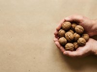 Caucasian hand holding a handful of walnuts in shells against a neutral beige background, representing healthy snacking, nutrition, and organic food selection.