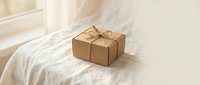 Kraft paper gift box with twine bow placed on white bedding near window with natural sunlight, suitable for holiday, birthday, and e-commerce packaging concepts.