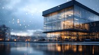 Modern glass office building exterior at dusk with snow falling, reflecting in still water. Warm interior lights create contrast against cool blue winter evening.