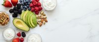 Fresh healthy breakfast ingredients with mixed berries, sliced avocado, nuts and yogurt arranged on white marble surface for nutritious meal preparation.