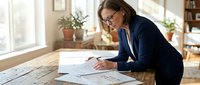 Professional businesswoman in navy blazer reviewing financial documents and charts at wooden desk in bright modern office with natural lighting.