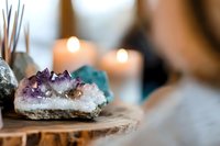 Raw amethyst crystal cluster on wooden surface with glowing candles and healing stones in soft focus background creating peaceful spiritual atmosphere.