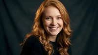 Professional headshot of smiling young Caucasian woman with curly red hair wearing black sweater against dark background for business portraits.